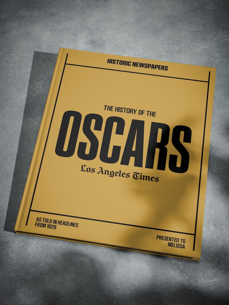 Puede incluir: Un libro amarillo con el t&iacute;tulo "The History of the Oscars" en letras negras. El libro es del Los Angeles Times y presenta titulares de 1929. El libro se presenta a Melissa.
