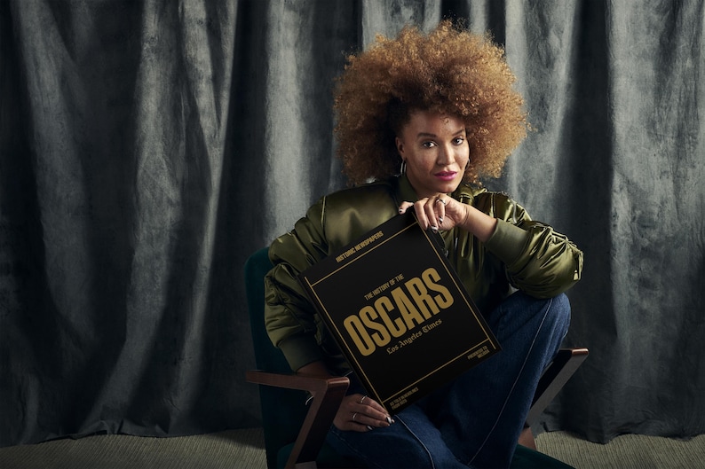 Puede incluir: Una mujer con un peinado afro est&aacute; sentada en una silla y sostiene un libro negro con letras doradas que dice "The History of the Oscars Los Angeles Times".