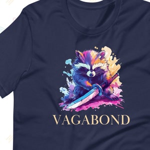 Pode incluir: Camiseta azul marinho com uma ilustração colorida de um guaxinim segurando uma espada, com a palavra "VAGABOND" em letras douradas abaixo. O guaxinim é retratado com azuis, roxos e amarelos vibrantes, em um fundo colorido.