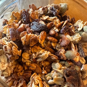May include: A close-up of a glass jar filled with granola. The granola is a mixture of oats, nuts, seeds, and dried fruit. The granola is a light brown color and has a slightly crunchy texture.