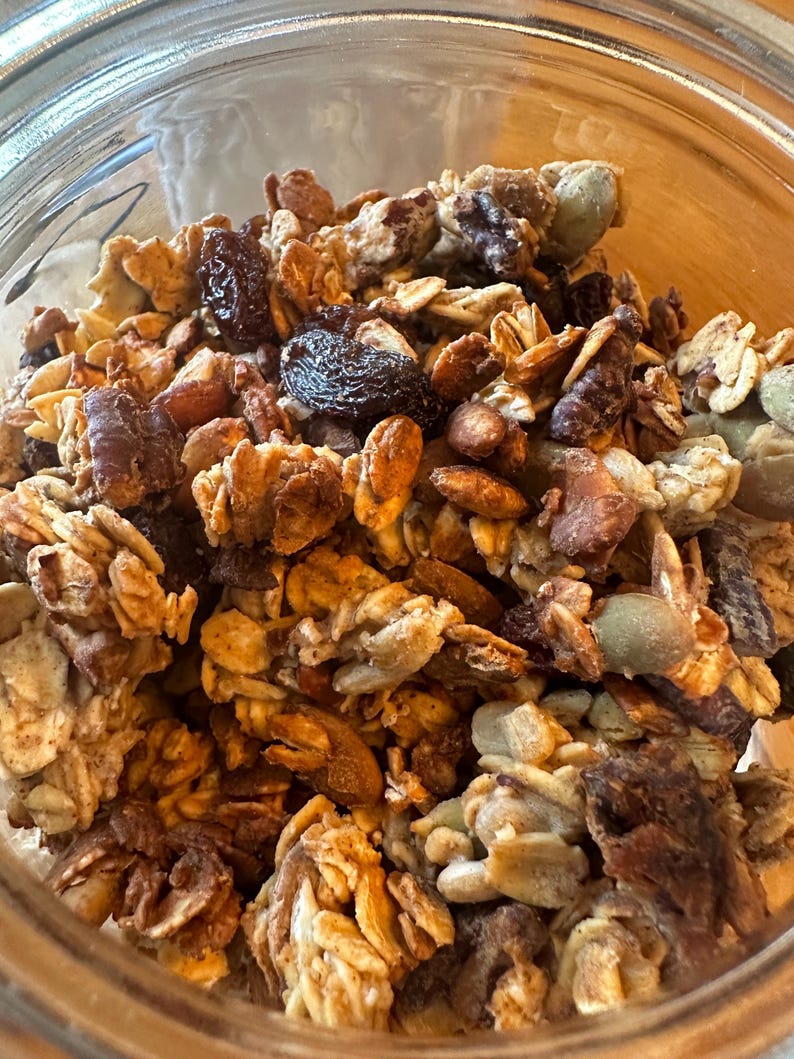 May include: A close-up of a glass jar filled with granola. The granola is a mixture of oats, nuts, seeds, and dried fruit. The granola is a light brown color and has a slightly crunchy texture.