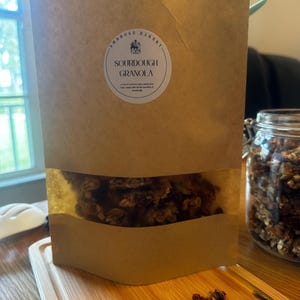 May include: A brown paper bag with a clear window showing sourdough granola inside. The bag has a white label with the text "Ambrose Bakery" and "Sourdough Granola".