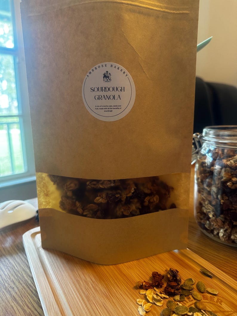 May include: A brown paper bag with a clear window showing sourdough granola inside. The bag has a white label with the text "Ambrose Bakery" and "Sourdough Granola".