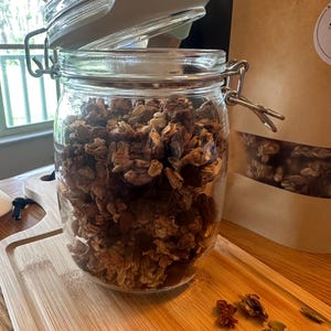 May include: A glass jar filled with granola, with the lid slightly ajar. The granola is a mixture of oats, nuts, seeds, and dried fruit. The jar is sitting on a wooden cutting board.