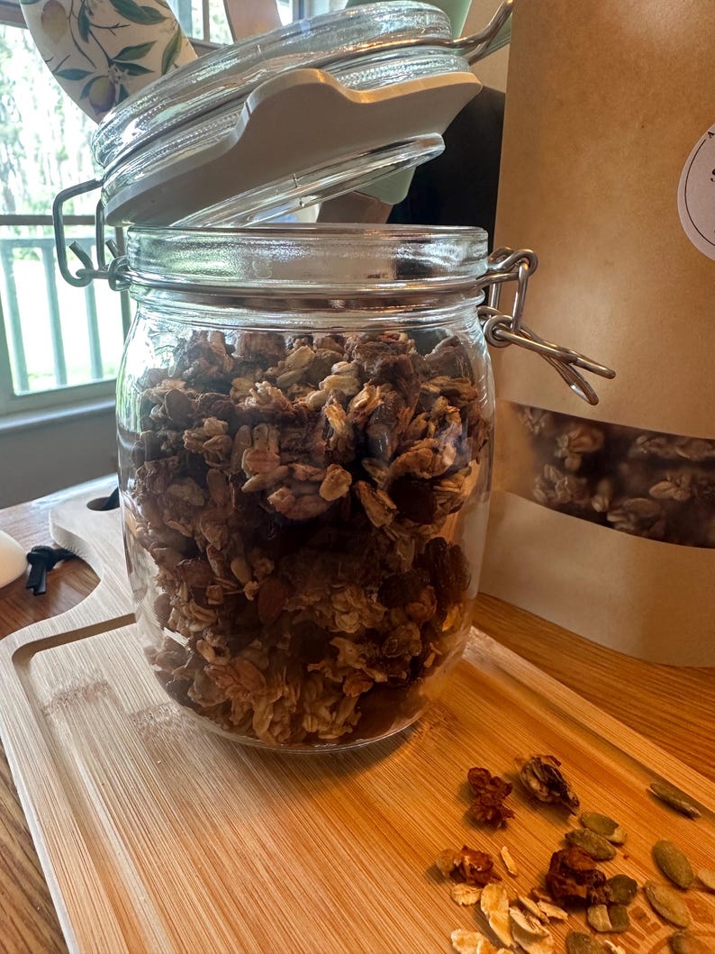 May include: A glass jar filled with granola, with the lid slightly ajar. The granola is a mixture of oats, nuts, seeds, and dried fruit. The jar is sitting on a wooden cutting board.