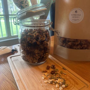 May include: A glass jar filled with sourdough granola, with the lid slightly ajar. The jar is sitting on a wooden cutting board with some granola scattered around it. A brown paper bag with the text "Sourdough Granola" is visible in the background.