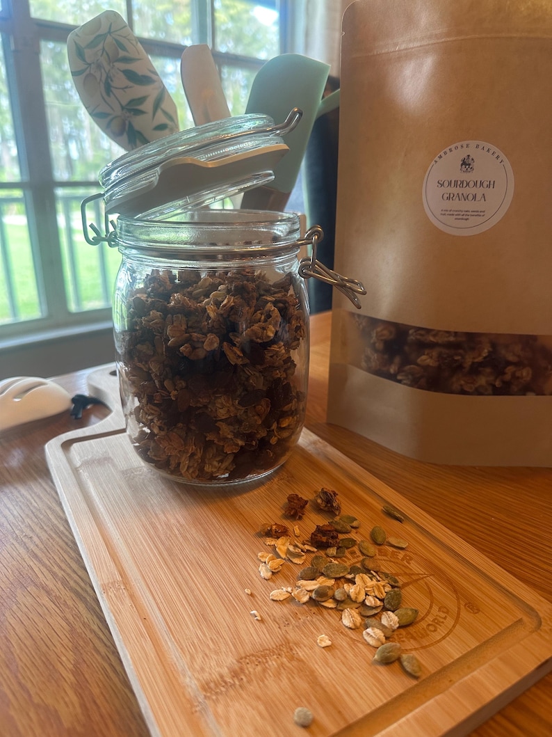 May include: A glass jar filled with sourdough granola, with the lid slightly ajar. The jar is sitting on a wooden cutting board with some granola scattered around it. A brown paper bag with the text "Sourdough Granola" is visible in the background.
