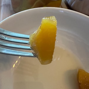 May include: A close-up of a fork holding a piece of preserved orange. The orange slice is a bright yellow-orange color and has a textured surface. A white bowl and a glass jar are in the background.