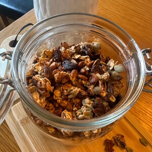May include: A glass jar filled with granola, a mixture of oats, nuts, seeds, and dried fruit. The granola is a light brown color and has a slightly crunchy texture.