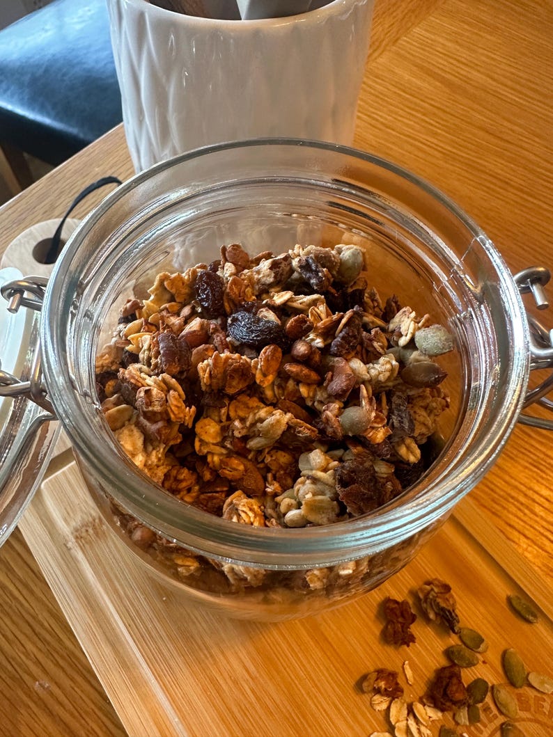 May include: A glass jar filled with granola, a mixture of oats, nuts, seeds, and dried fruit. The granola is a light brown color and has a slightly crunchy texture.