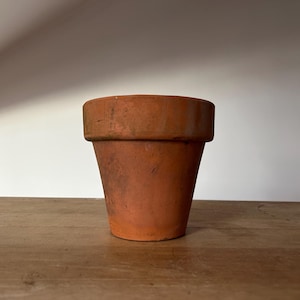 May include: A terracotta flower pot with a slightly weathered appearance. The pot is empty and sitting on a wooden surface.