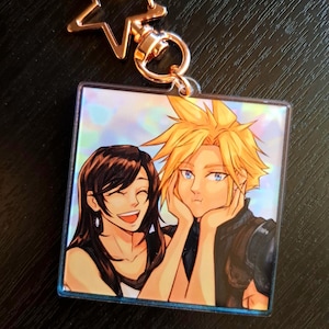 May include: A holographic keychain featuring a cartoon image of a man and woman with blonde and brown hair. The man is smiling and the woman is laughing. The keychain is attached to a gold star-shaped charm.