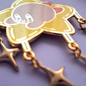 May include: A gold-colored enamel pin featuring a cartoon character in a yellow star shape with two dangling star charms. The character has a pink ear, blue eyes, and a light gray body.