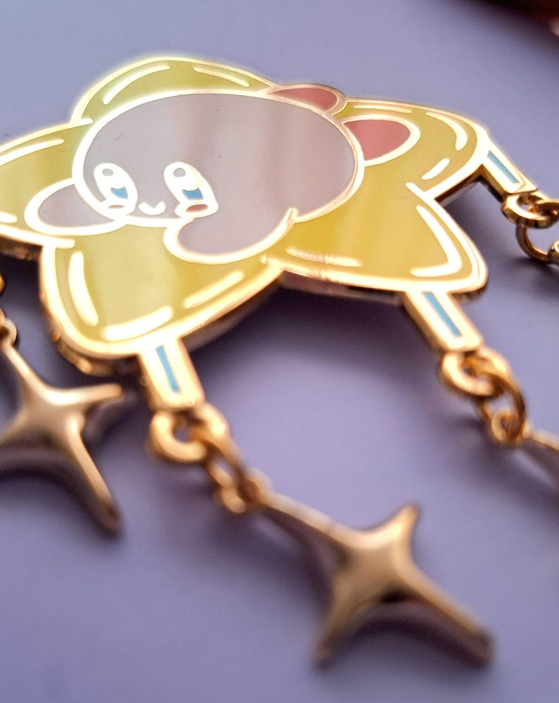 May include: A gold-colored enamel pin featuring a cartoon character in a yellow star shape with two dangling star charms. The character has a pink ear, blue eyes, and a light gray body.