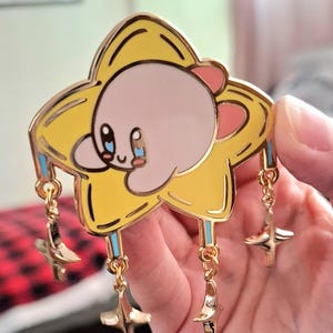 May include: A gold-colored enamel pin featuring a pink Kirby character holding onto a yellow star with blue and white accents. The star has four smaller gold stars dangling from it.