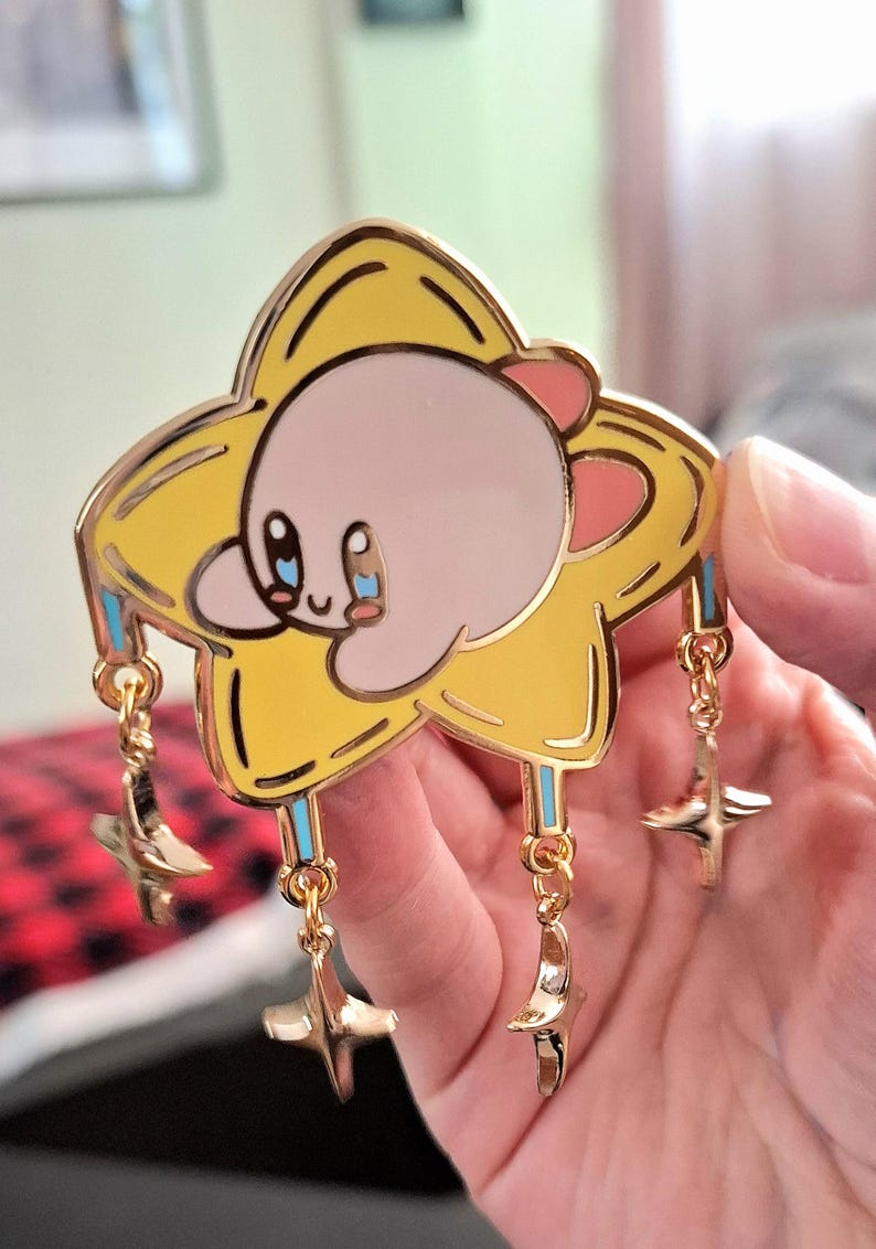 May include: A gold-colored enamel pin featuring a pink Kirby character holding onto a yellow star with blue and white accents. The star has four smaller gold stars dangling from it.