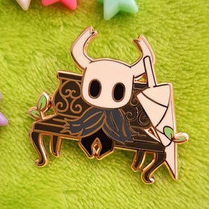 以下が含まれることがあります： ビデオゲーム「Hollow Knight」のキャラクターが装飾的なベンチに座っている、金色のエナメルピン。ピンは白い顔、黒い目、濃い青色のマントをしています。白と緑の傘が横に持たれています。ピンク、緑、白の星型のビーズが背景にあります。