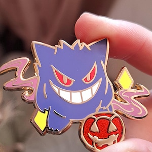 Puede incluir: Pin de esmalte dorado con un Gengar Pokémon morado y azul sosteniendo una calabaza roja. Gengar tiene una sonrisa blanca y ojos rojos. El pin tiene un efecto de humo rosa y amarillo.