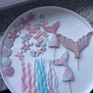 Könnte beinhalten: Ein Set aus lila, rosa und blauen Fondant-Meerjungfrauen-Kuchen-Toppern. Das Set enthält zwei Meerjungfrauen-Schwänze, zwei Meerjungfrauen-Oberteile, Muscheln, Seesterne und Seetang.