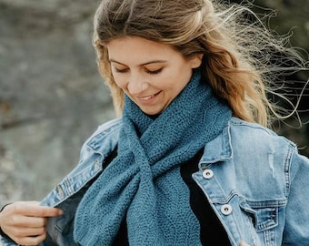 Puede incluir: Una bufanda de punto azul verdoso se usa sobre una camiseta negra y una chaqueta vaquera. La bufanda está envuelta alrededor del cuello y cruzada por delante. La bufanda tiene un patrón texturizado.