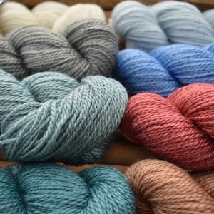 May include: A collection of yarn skeins in various shades of blue, grey, red, brown, and white. The yarn is arranged in a row, with each skein overlapping the next.