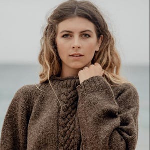 Könnte beinhalten: Ein brauner Strickpullover mit Zopfmuster und hohem Kragen. Der Pullover ist aus weichem, warmem Material gefertigt und hat eine lockere Passform.