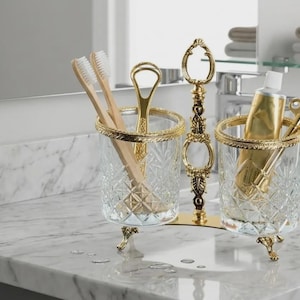 May include: A decorative bathroom organizer with two crystal glasses, each with a gold rim and ornate gold feet. One glass holds toothbrushes, the other holds a tube of toothpaste and a key. A gold stand connects the glasses.