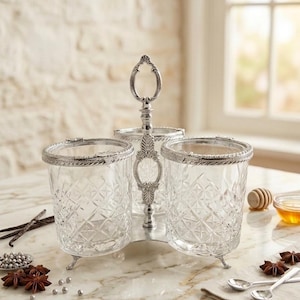 May include: A three-section serving dish with a silver-colored handle and trim. The clear glass containers have a diamond pattern. The dish sits on a marble-like surface, with spices and small spoons nearby. A decorative piece for serving condiments.
