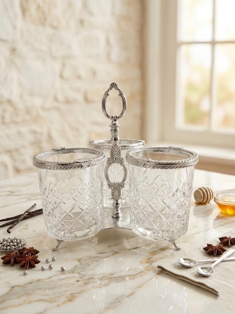 May include: A three-section serving dish with a silver-colored handle and trim. The clear glass containers have a diamond pattern. The dish sits on a marble-like surface, with spices and small spoons nearby. A decorative piece for serving condiments.