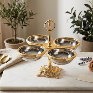 May include: A gold-colored, four-tiered serving dish with ornate detailing. The dish has four stainless steel bowls with gold rims, connected by a central stem. The base is square and also gold. The dish sits on a white marble surface.