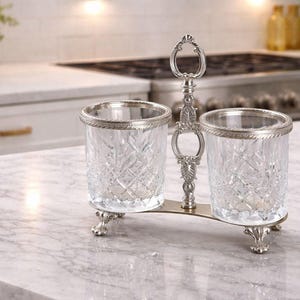 May include: A silver-toned condiment holder with two cut-glass containers. The containers have a diamond pattern and are trimmed with silver. The holder has a decorative handle and rests on ornate feet. The background shows a kitchen countertop.