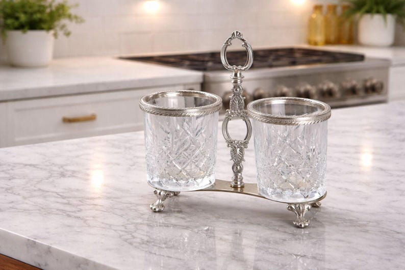May include: A silver-toned condiment holder with two cut-glass containers. The containers have a diamond pattern and are trimmed with silver. The holder has a decorative handle and rests on ornate feet. The background shows a kitchen countertop.