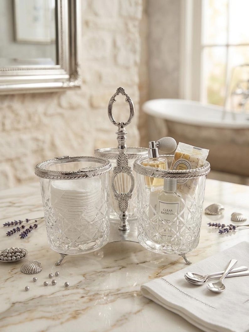 May include: A three-sectioned, silver-toned vanity organizer with clear glass containers. One section holds cotton pads, another has a perfume bottle and bath oil, and the third has cotton swabs. The organizer sits on a marble surface.