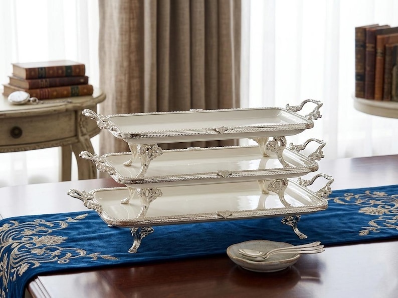 Op de afbeelding: Een drielaagse serveerschaalset met een wit oppervlak en sierlijke zilverkleurige rand en handgrepen. De schalen worden ondersteund door decoratieve poten en liggen op een blauwe geborduurde loper. Een klein schaaltje met lepels staat ervoor.