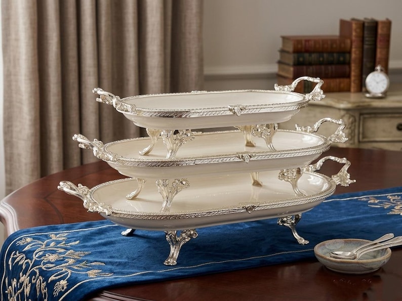 Op de afbeelding: Een serveerschaal met drie niveaus, met een cr&egrave;mekleurig oppervlak en zilverkleurige randen en handgrepen. De schalen zijn ovaal van vorm en worden ondersteund door sierlijke zilverkleurige poten. De schaal staat op een donkere houten tafel met een blauwe loper.