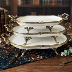Può includere: Vassoi da portata a tre livelli con superfici in ceramica bianca e finiture e manici in metallo color oro ornate. I vassoi sono di forma ovale e poggiano su gambe decorative. Lo sfondo comprende libri antichi e un runner blu petrolio scuro.