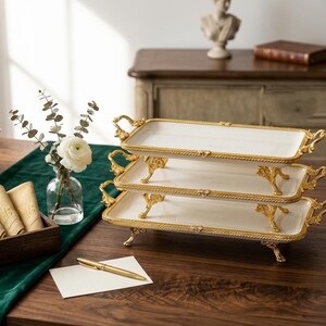Op de afbeelding: Dienbladen met drie niveaus met witte oppervlakken en goudkleurige randen en handgrepen. De rechthoekige trays staan op sierlijke goudkleurige poten. Een gouden pen en een blanco kaart liggen op tafel.