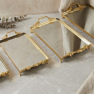 Pode incluir: Quatro bandejas decorativas retangulares com superfícies espelhadas e molduras douradas ornamentadas. Cada bandeja tem uma alça e pés decorativos. As bandejas estão dispostas sobre uma superfície semelhante a mármore, com um vaso de flores ao fundo.