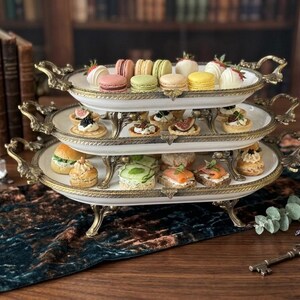 Op de afbeelding: Een serveerschaal met drie niveaus, met een goudkleurig metalen frame en witte porseleinen borden. De schalen zijn gevuld met een assortiment kleurrijke macarons, miniatuursandwiches en hapjes. De achtergrond omvat antieke boeken en een donkergroene tafelloper.