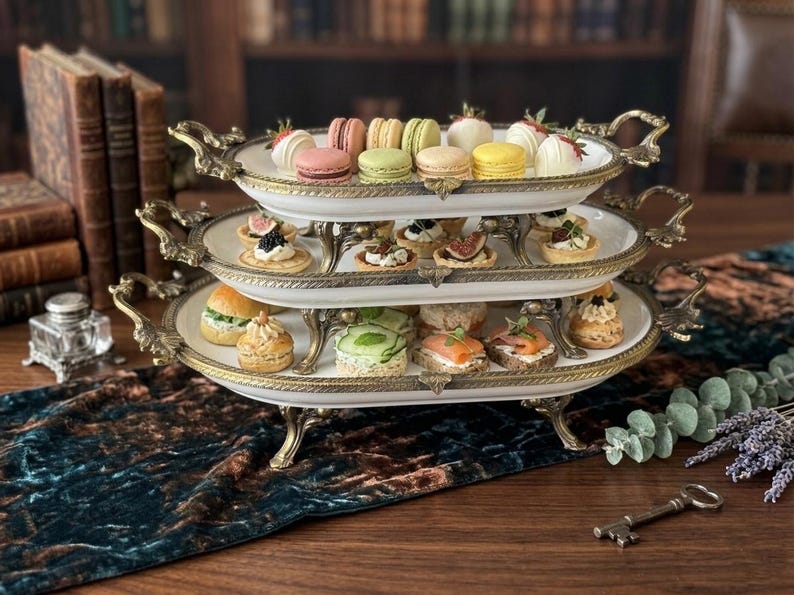 Op de afbeelding: Een serveerschaal met drie niveaus, met een goudkleurig metalen frame en witte porseleinen borden. De schalen zijn gevuld met een assortiment kleurrijke macarons, miniatuursandwiches en hapjes. De achtergrond omvat antieke boeken en een donkergroene tafelloper.