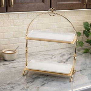 Può includere: Un alzatina a due livelli con struttura in metallo color oro e due piatti rettangolari in ceramica bianca. L'alzatina ha un manico ad arco nella parte superiore ed è appoggiata su un piano di lavoro in marmo. Stoviglie decorative sullo sfondo.