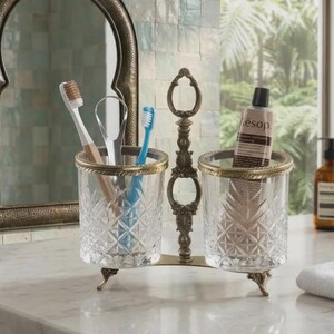 May include: A decorative toothbrush and toothpaste holder with two clear glass cups, each with a gold rim and diamond pattern. The holder has a bronze-colored metal frame and is on a marble countertop. The image also includes a mirror and a tube of Aesop toothpaste.