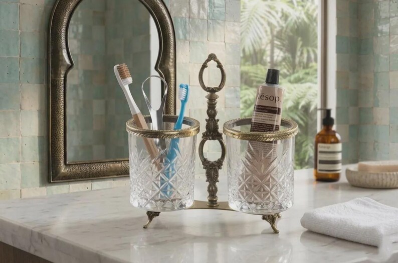 May include: A decorative toothbrush and toothpaste holder with two clear glass cups, each with a gold rim and diamond pattern. The holder has a bronze-colored metal frame and is on a marble countertop. The image also includes a mirror and a tube of Aesop toothpaste.