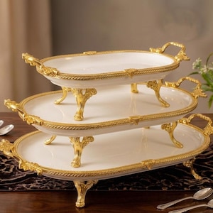 May include: A three-tiered serving tray with a white surface and gold trim. The tray has ornate gold handles and legs. The trays are oval-shaped and are stacked on top of each other. The tray is on a dark brown table.