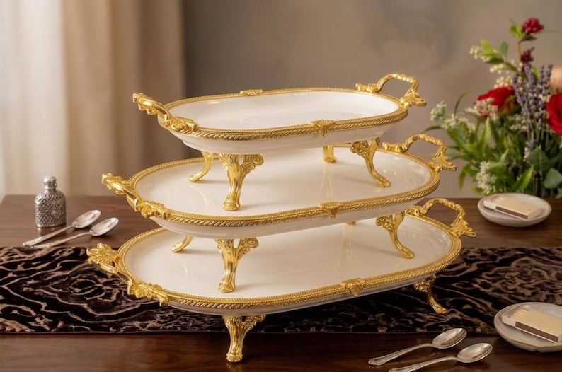Op de afbeelding: Een serveerschaal met drie niveaus met een wit oppervlak en gouden rand. De schaal heeft sierlijke gouden handvatten en poten. De schalen zijn ovaal van vorm en zijn op elkaar gestapeld. De schaal staat op een donkerbruine tafel.
