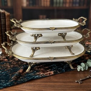 Op de afbeelding: Een set van drie serveerschalen met ovale witte porseleinen schalen. Elke schaal heeft een decoratieve goudkleurige metalen rand, handgrepen en sierlijke poten. De schalen zijn op elkaar gestapeld, wat een aantrekkelijke presentatie cre&euml;ert.