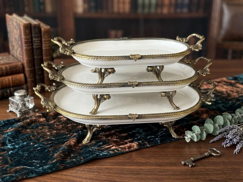 Op de afbeelding: Een set van drie serveerschalen met ovale witte porseleinen schalen. Elke schaal heeft een decoratieve goudkleurige metalen rand, handgrepen en sierlijke poten. De schalen zijn op elkaar gestapeld, wat een aantrekkelijke presentatie cre&euml;ert.