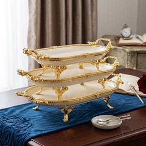 Op de afbeelding: Een serveerschaal met drie niveaus, met een cr&egrave;mekleurig oppervlak en gouden randen. De ovale schalen worden ondersteund door sierlijke gouden poten. De handgrepen zijn ook goudkleurig. De schalen zijn gestapeld op een donkere houten tafel met een blauwe loper.