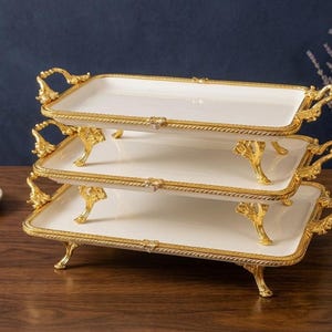 Op de afbeelding: Een serveerschaal met drie niveaus met een wit oppervlak en goudkleurige randen en handgrepen. De schaal heeft sierlijke gouden poten en staat op een houten oppervlak. Een klein schaaltje met zilver verpakte snoepjes staat op de achtergrond.