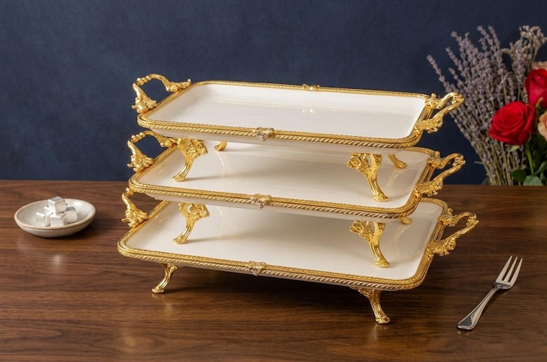 Op de afbeelding: Een serveerschaal met drie niveaus met een wit oppervlak en goudkleurige randen en handgrepen. De schaal heeft sierlijke gouden poten en staat op een houten oppervlak. Een klein schaaltje met zilver verpakte snoepjes staat op de achtergrond.